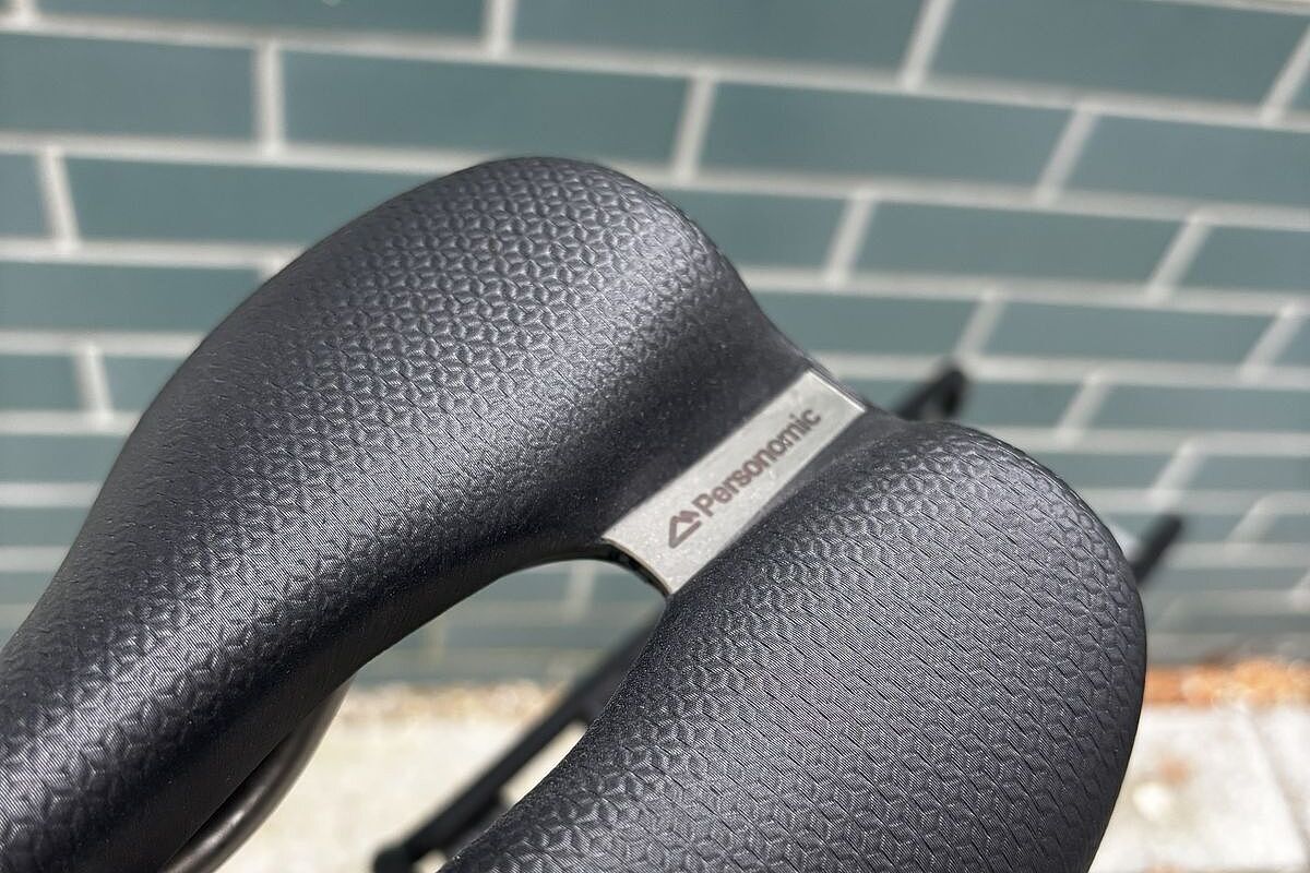 Detail eines schwarzen Fahrradsattels mit feiner Struktur und Metallplakette vor hellgrüner Kachelwand.