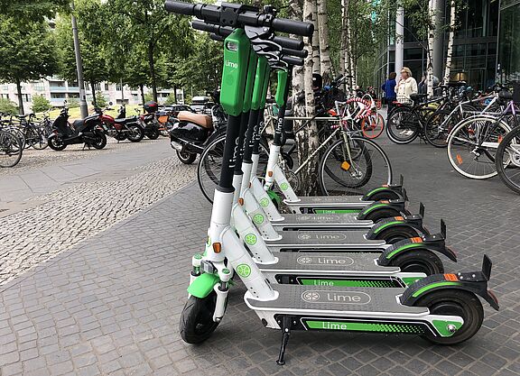 E-Scooter zum Ausleihen in Berlin Im Vordergrund mehrere E-Scooter eines Verleihs, im Hintergrund mehrere Fahrräder, einige Personen und Gebäude