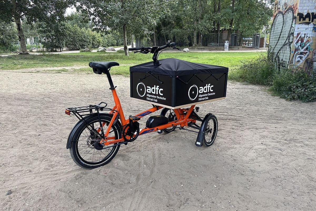 Orangefarbenes dreirädriges Lastenrad mit schwarzer ADFC-Transportbox steht auf einem sandigen Weg im Grünen.