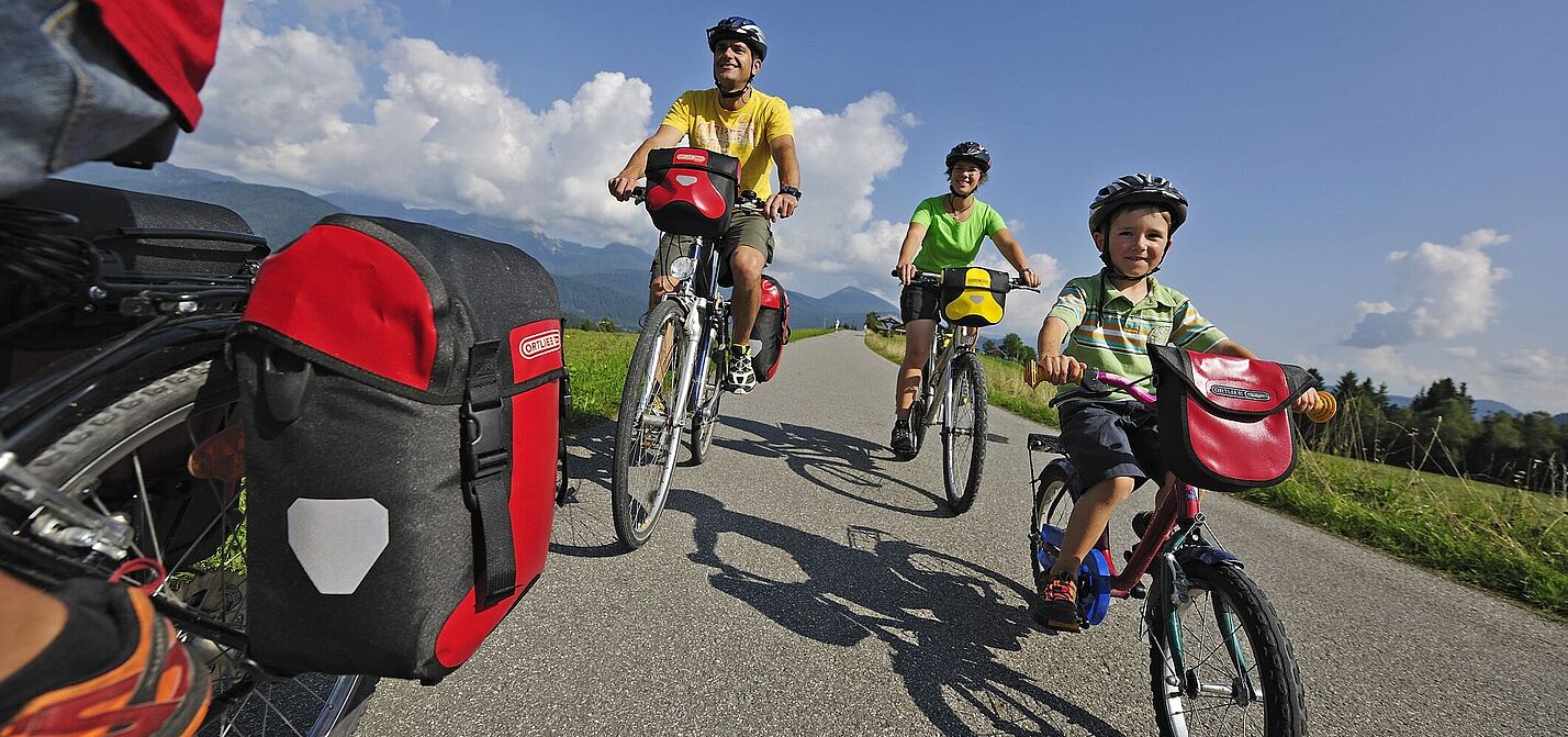 Sommerliche Familienradtour auf dem Land Eine vierköpfige Familie fährt mit Helmen und Packtaschen auf Fahrrädern eine schmale Landstraße entlang; vorn ein Kind auf einem kleinen Rad, dahinter zwei Erwachsene und ein weiteres Fahrrad im Anschnitt links, umgeben von Wiesen, Bergen und blauem Himmel.